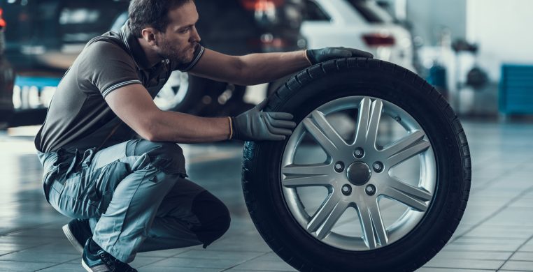 Caucasian Handsome Auto Mechanic Cheking Car Wheel. Portrait of Adult Machinist Doing Wearing Uniform Work in Garage. Technic occupation. Automobile Repair Service Concept.